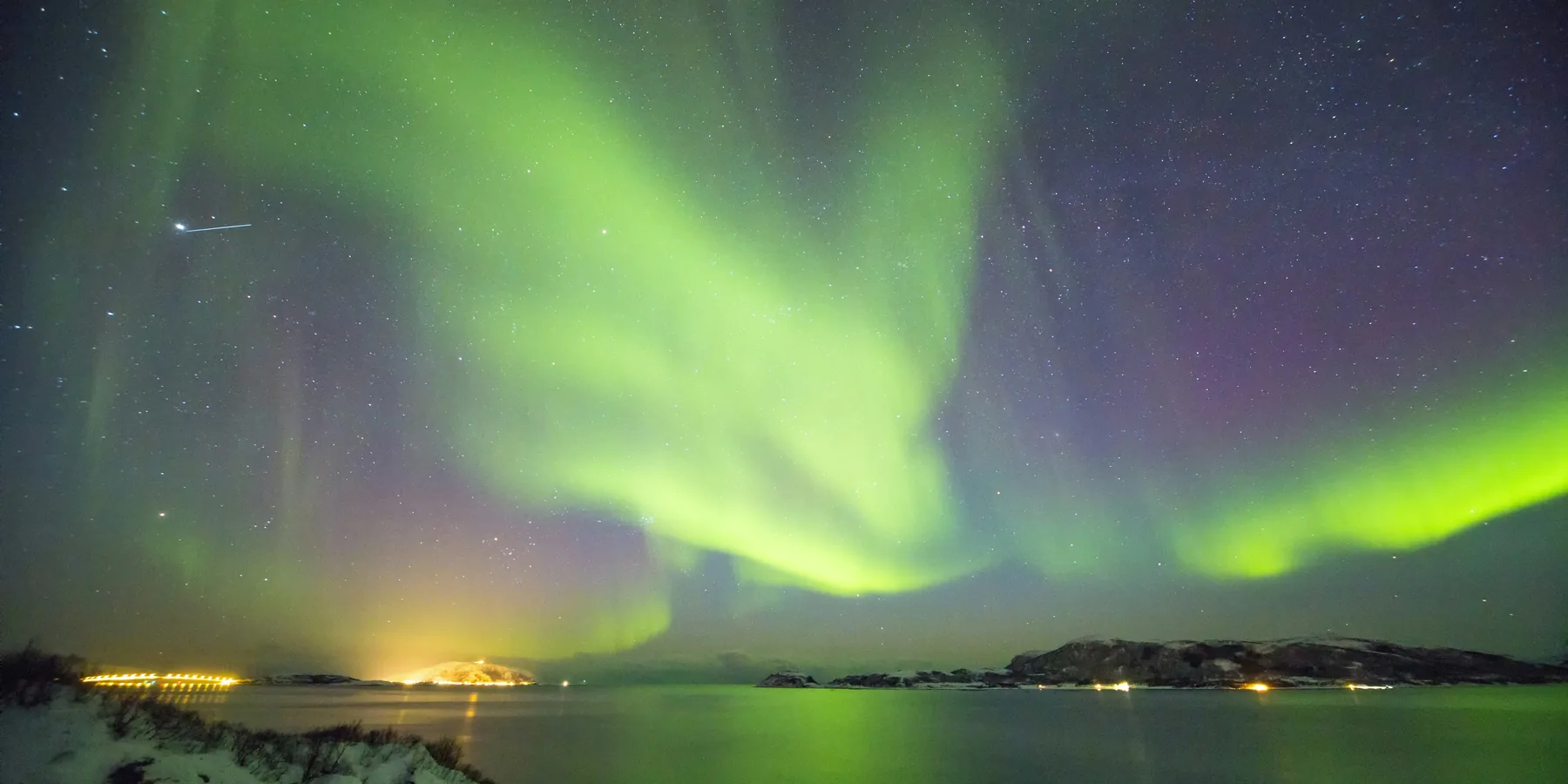 Northern lights in green and violet over dark water and the lights of the shore near Tromsø
