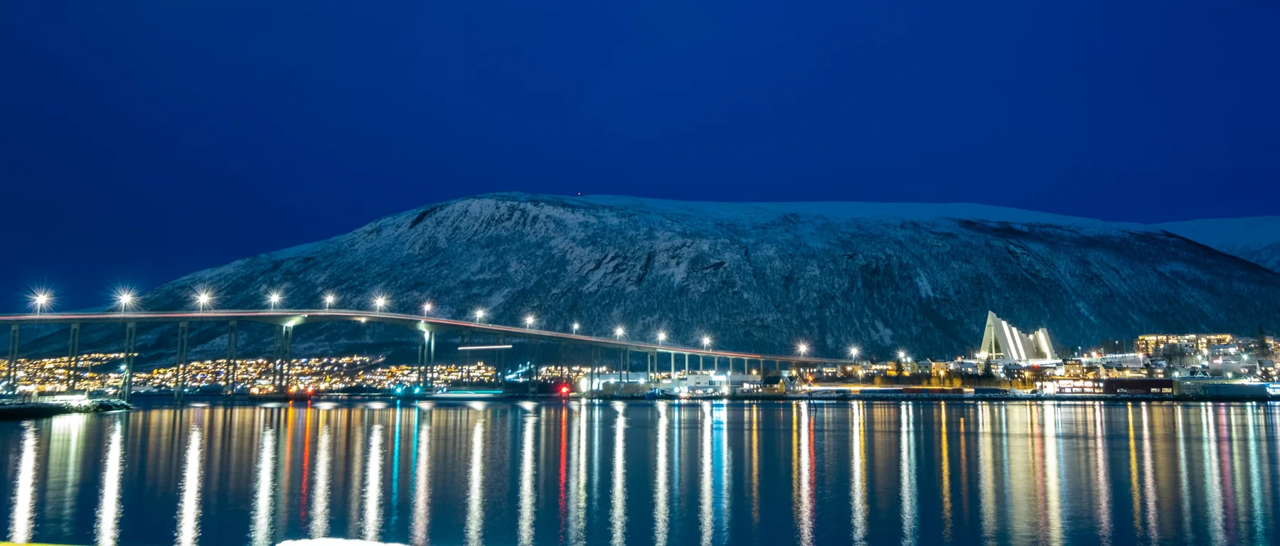 The lit Tromsø Bridge and the Arctic Cathedral below a snowy mountain, reflected in the strait at blue hour