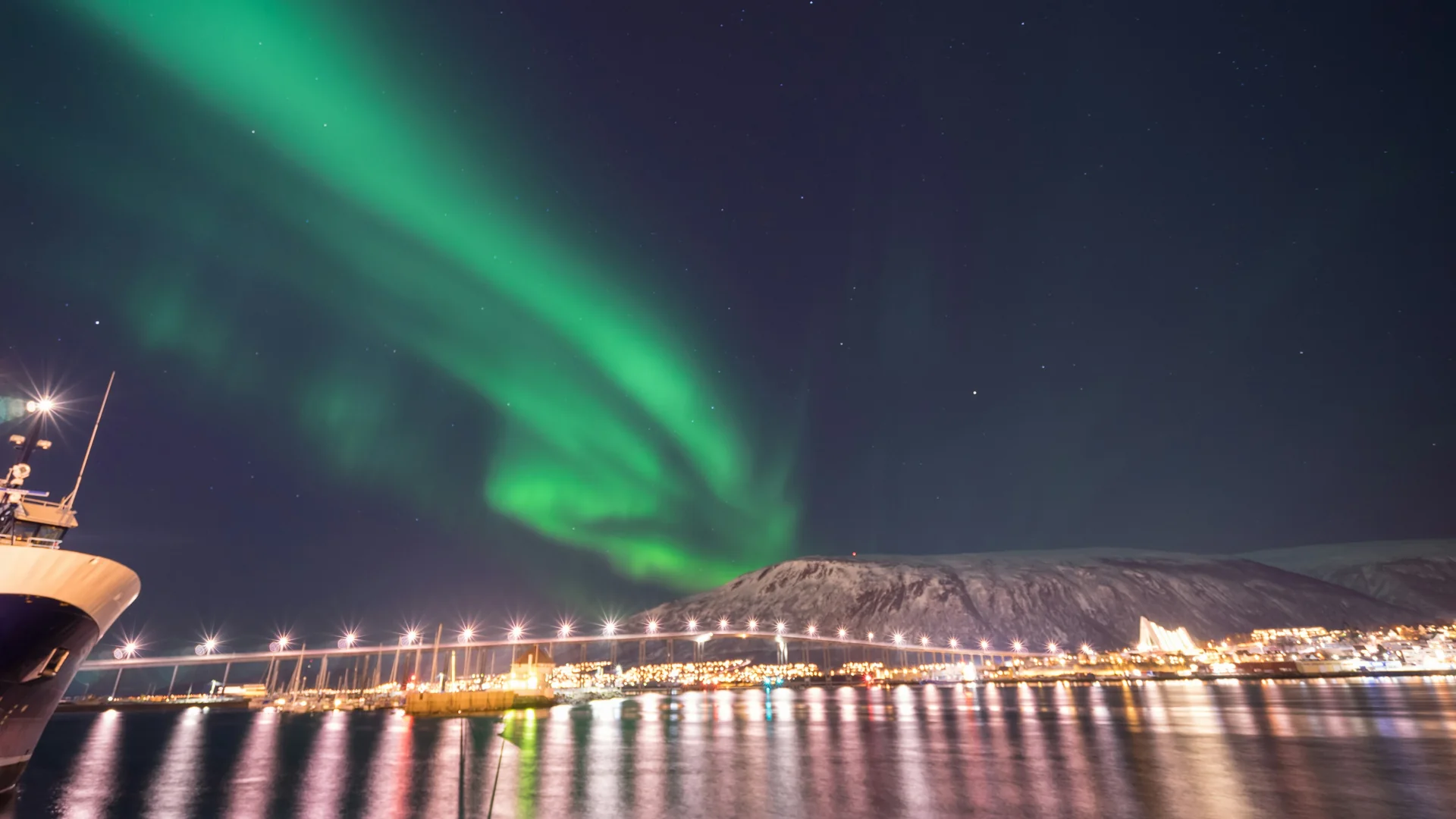 Green northern lights arching over the lit Tromsø Bridge and the Arctic Cathedral, seen across the harbour at night